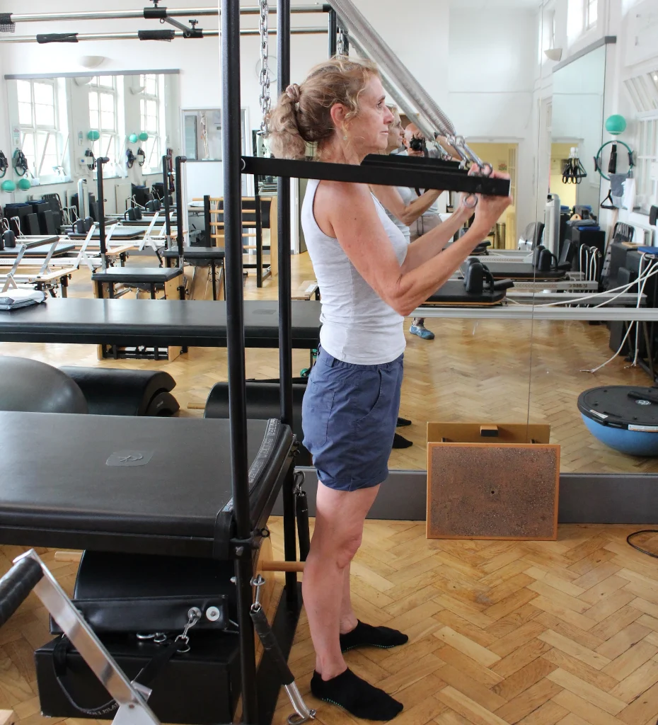 A client in her sixties standing tall at the Cadillac, drawing the roll-down bar towards her chest, at Applied Pilates, London Colney, St Albans